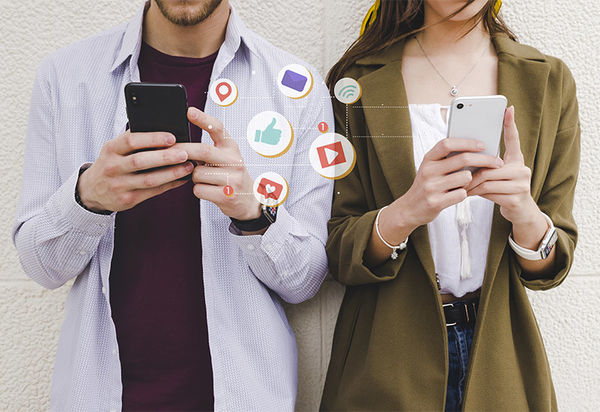 Zwei junge Menschen, ein Mann und eine Frau, stehen nebeneinander und halten jeweils ihr Mobiltelefon in der Hand. Zwischen den Telefonen schweben die Symbole verschiedener sozialer Netzwerke wie facebook, twitter, instagram, youtube und andere.