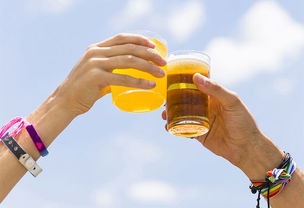 Zwei Hände stoßen vor dem blauen Himmel im Hintergrund, die Gläser, die sie halten zusammen. Beide tragen Festivalbändchen aber auch Freundschaftsbänder.