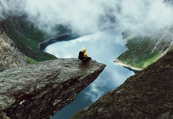Am Rand einer ins Bild hinein ragenden Felsklippe sitzt eine Person und sieht in den vor ihr liegenden Abgrund und auf den See unter ihr, der sich zwischen Berghängen aufspannt. Die Gipfel der den See umgebenden Berge, von denen sich einige durch die Wolken abzeichnen sind schneebedeckt. Die Person in der gelben Jacke mit dem schwarzen Rucksack auf dem Rücken scheint allein zu sein in dieser Weite. Auch wie sie sich fühlt ist nicht erkennbar. Groß, da sie über allem thront, oder klein, angesichts dieser Naturhaften Gewalt, die so ohne jegliches Zutun des Menschen einfach so da ist?