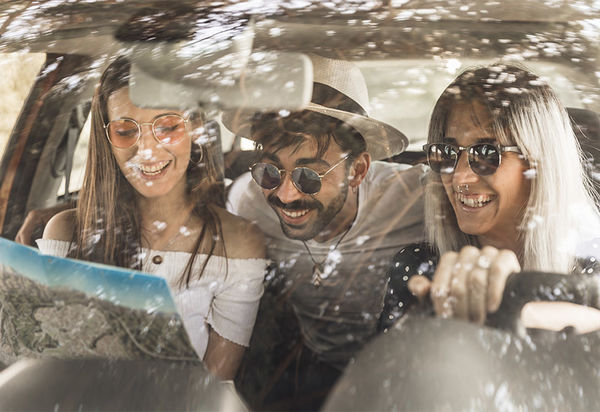 Wir blicken durch die Windschutzscheibe in das Innere eines Autos, in dem drei junge Leute sitzen. Sie tragen Sonnenbrillen und lächeln oder lachen alle. Die Beifahrerin auf der rechten Seite guckt in eine Karte. Der junge Mann mit Hut, der auf der Rückbank sitzt, beugt sich zu ihr nach vorne und sieht über ihre Schulter ebenfalls in die Karte. Ebenso, wie die junge Frau mit Nasenpiercing, die am Steuer sitzt.