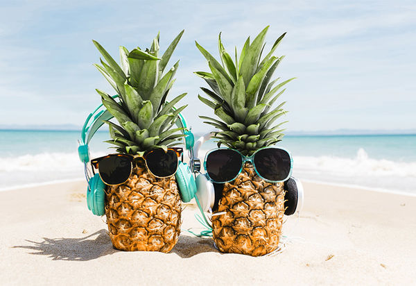 Zwei Ananas mit Sonnenbrillen stehen im weißen Sand an einem Strand. Im Hintergund sieht man türkises, welliges Meer. Beide haben als Accessoire neben den Sonnenbrillen auch noch Kopfhörer auf. 