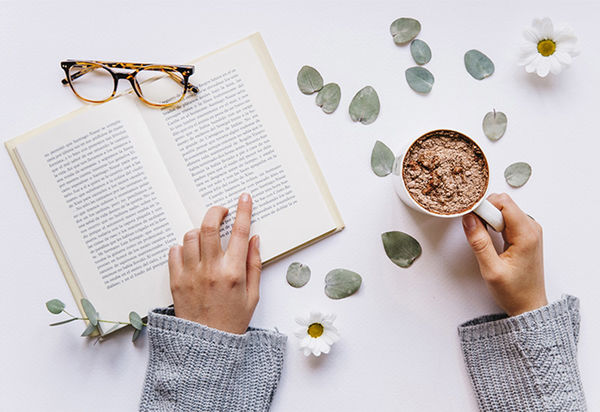 Von oben blickt man auf ein aufgeschlagenes Buch in dem eine menschliche Hand auf eine Zeile zeigt, so als ob sie mit dem Finger beim Lesen den Text entlang fährt. Die andere dazugehörige Hand hält eine Tasse mit einem bräunlichen Getränk. Vermutlich handelt es sich dabei um Kakao. Auf dem Buch liegt noch eine Brille. Einige Eukalyptusblätter und Margeritenblüten sind um das Buch und die Hände verstreut. Es sieht nach einem gemütlichen Lesenachmittag mit Lieblingskakao aus.  