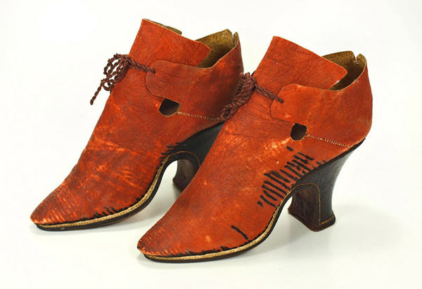 Damenstöckelschuh aus rotem Schafleder mit Resten von Verzierungen aus schwarzen Borten am gesamten Schaft. Die Stepplöcher sind noch sichtbar. Das Vorderteil endet nach oben in großer eckiger Ristlasche. Als Verschluss sind zwei kurze Fersenteillaschen, die mittels eines Bandes über der Ristlasche zusammengebunden wurden. Über dem Sohlenrand ist eine weiße Lederpaspel eingearbeitet. Absatz leicht geschweift, mit schwarzem Leder überzogen und Halbmond-Trittflächen. Sohlen- und Absatzlinie werden durch eine weiße Naht markiert.