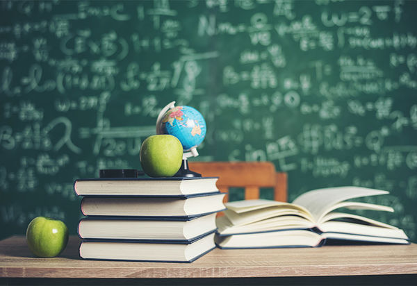 Im Hintergrund eine Schultafel, die in weißer Kreideschrift von oben bis unten mit mathematischen Formeln beschriftet ist. Vor der Tafel im Hintergrund steht ein Tisch mit einem etwas abgerückten Stuhl. Auf diesem Tisch, auf den wir von vorne Blicken liegen einige Bücher  auf zwei Stapeln. Auf dem höheren liegt ein Apfel neben einem kleinen Globus und einer Lupe? Auf dem anderen, kleineren Stapel liegt ein aufgeschlagenes Buch. Ein weiterer Apfel befindet sich links des größeren Stapels. Es hat den Anschein, als ob dort bis eben jemand gesessen und gelernt hätte.