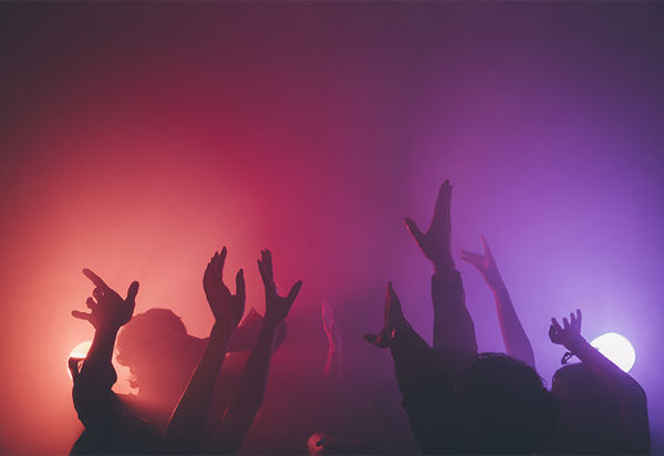 In rosa-lila-farbigem Licht recken sich zahlreiche Arme von Tanzenden in die Höhe. Im Hintergrund sieht man zwei helle Scheinwerfer. Man kann sich beim Betrachten des Bildes gut vorstellen, dass die Musik dort ziemlich laut ist und die Leute viel Spass beim Tanzen haben.