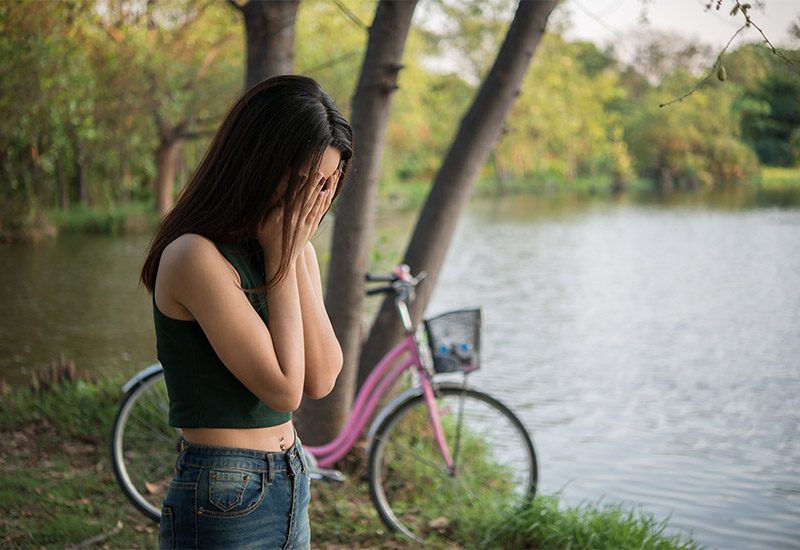 Eine junge Frau steht weinend an einem Waldsee. Sie hält sich die Hände vor das Gesicht. Im Hintergrund lehnt ihr Fahrrad an einem Baum.
